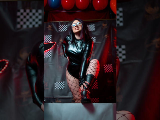 CristinaSmit in a shiny black leather bodysuit, fishnet stockings, and black boots, wearing a blue and white eye mask, smiling with red lights in the background. Boobs, ass, pussy.