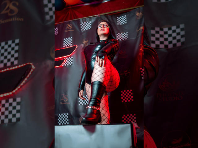 CristinaSmit in black latex dress, fishnet stockings, and knee-high boots, standing with hand on thigh, red light on left, wearing glasses, confident pose, sexy, dark background.