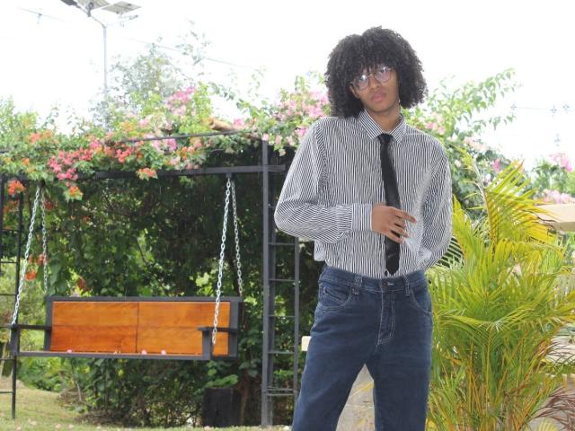 AntonyCarterr with curly black hair, glasses, striped shirt, black tie, and blue jeans, standing next to a wooden swing with metal chains.