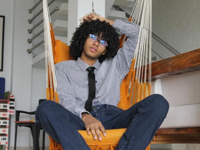 AntonyCarterr with curly black hair, glasses, striped shirt, black tie, and blue jeans, sitting in an orange hammock, legs spread, hand in hair, looking relaxed.
