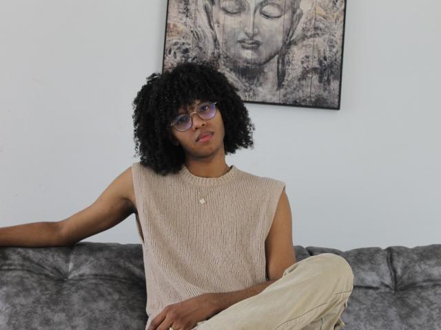 AntonyCarterr with curly black hair, glasses, sleeveless beige knit shirt, beige pants, sitting on gray couch, arms relaxed, looking slightly to the side, calm expression.