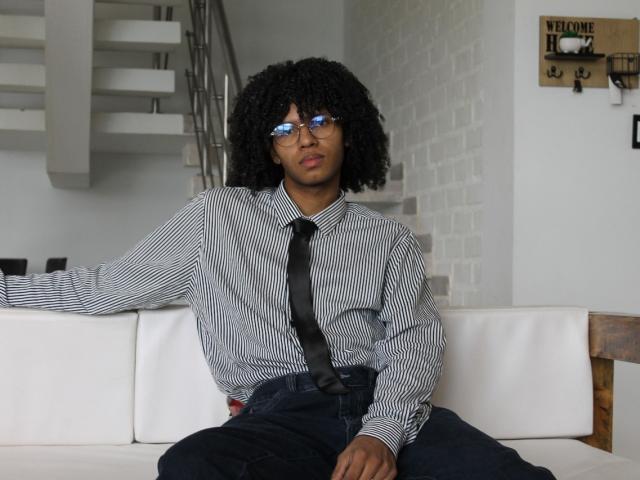 AntonyCarterr with curly black hair, blue glasses, striped shirt, black tie, and dark pants, sitting on white couch, looking straight ahead with a neutral expression.