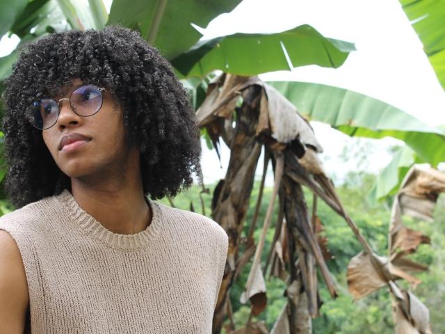 AntonyCarterr with curly black hair, wearing glasses and a beige knit sleeveless top, standing in front of green banana leaves and a brown, withered banana plant.