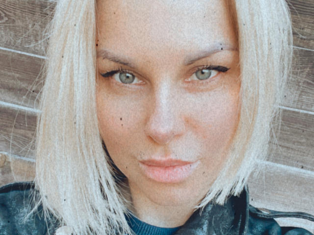 BelleLisaG has blonde hair, green eyes, black eyeliner, and a few dark moles on her face. She's wearing a black jacket with a blue shirt underneath.