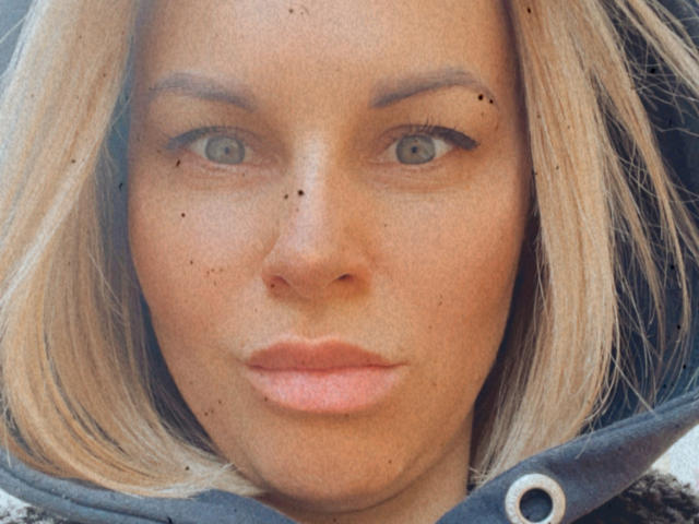 Het gezicht van BelleLisaG, met blond haar, blauwe ogen en een paar zwarte sproeten op haar neus en wangen, gekleed in een donkere hooded jas, die recht in de camera kijkt..