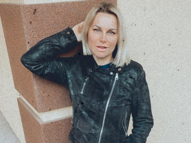 BelleLisaG with blonde bob, green eyes, black leather jacket, hand in pocket, leaning against red stone wall, slight smile, casual pose.