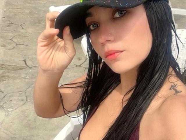 BrihannaSex with wet black hair, wearing a black cap, holding it, showing her nose ring, tattoo on right shoulder, red lips, and a deep cleavage in a pink top.
