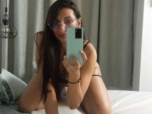 CelestePoot with long brown hair, glasses, and black lingerie, kneeling on a bed, taking a mirror selfie with a green phone, showing her tits and ass.