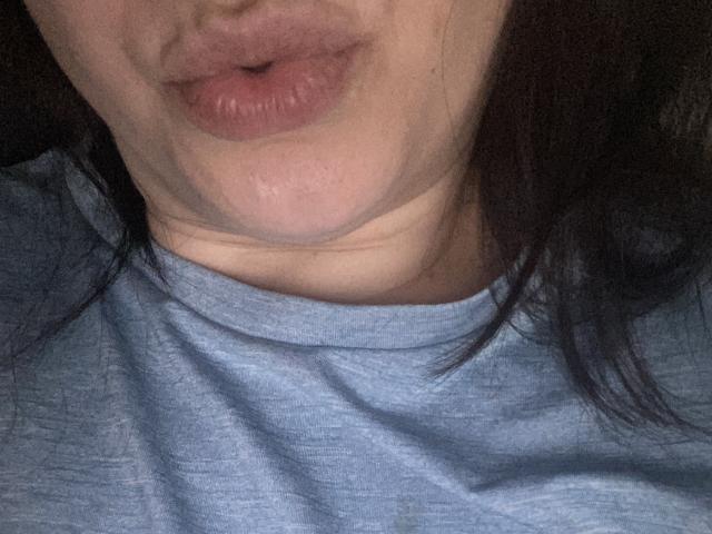 Los labios hinchados y la piel ligeramente flácida de JessicaJess69 son visibles en un selfie con poca luz. Lleva una camisa gris, y se puede ver parte de su cabello oscuro en el lado derecho..