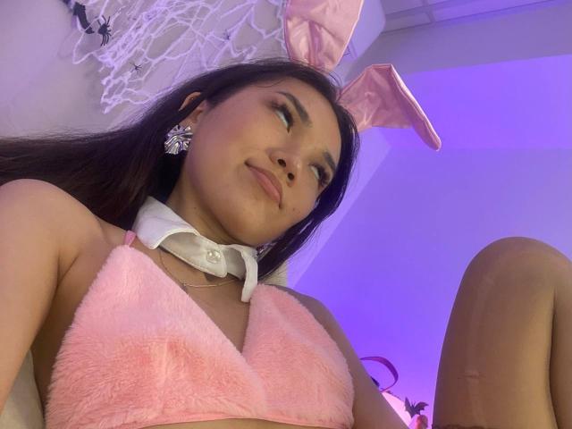DivineMadness with pink bunny ears, fluffy pink bra, white collar, and flower earrings, looking up with a slight smile, showing her tits and a cute, confident face.
