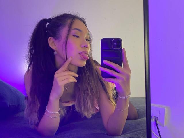 DivineMadness with long brown hair in pigtails, tongue out, finger near mouth, taking selfie in mirror, purple light, wearing bracelet, black top, holding black phone.