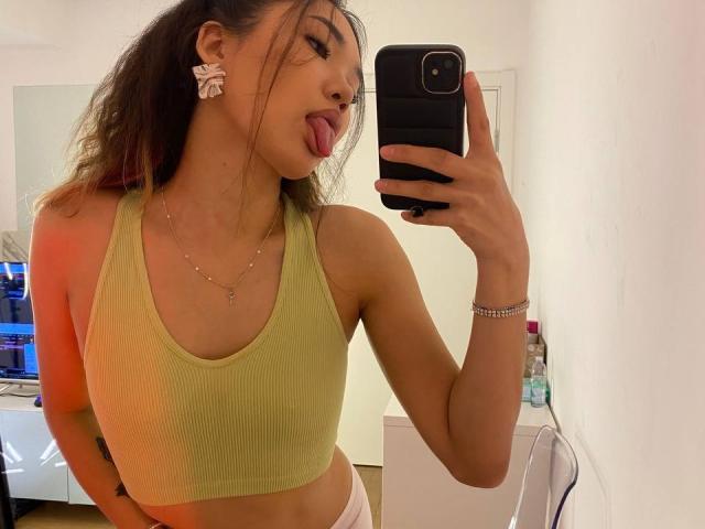 DivineMadness, Asian woman, green sports bra, white panties, sticking out tongue, long brown hair, ear clip, gold necklace, holding phone, mirror selfie, small tits, slim, bedroom.