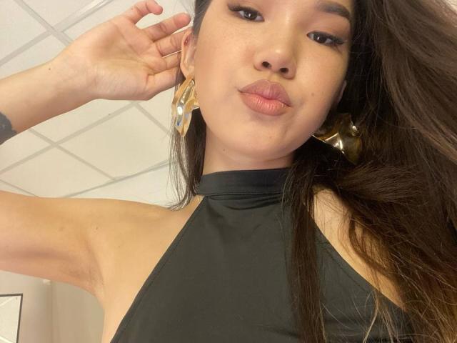 DivineMadness is a young woman with long, straight brown hair and medium brown skin. She's wearing a black halter top and gold geometric earrings. Her right hand is raised, touching her hair. She has full lips, slightly pouted, and subtle makeup with dark eyeliner. Her eyebrows are dark and slightly arched. The background is a tiled wall, slightly blurred. The image has a casual, selfie-style composition.