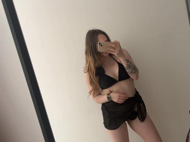 DakotaGold in a black bikini top with flower, showing her tits, and a black skirt, holding a phone, with long brown hair, tattoo on right arm, and a watch.