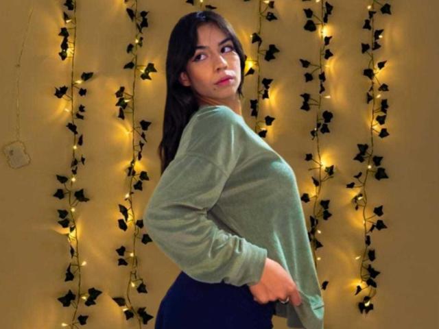 KatherinJohns with dark hair, green sweatshirt, and blue pants, looking back over her shoulder, standing in front of yellow fairy lights and black leaf decorations.