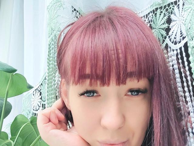 Kohka with pinkish-red hair, bangs, and black eyeliner, looking at camera, hand near face, green lace curtain, and green plant in background. Her eyes are blue and she's smiling.