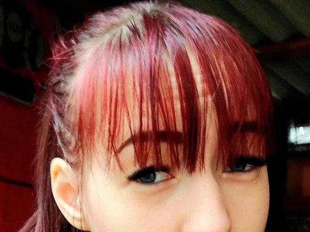 Kohka's face with red, pinkish-dyed hair, straight bangs, and blue eyes, wearing black eyeliner. Her nose is slightly visible, and she has a subtle, confident smile.