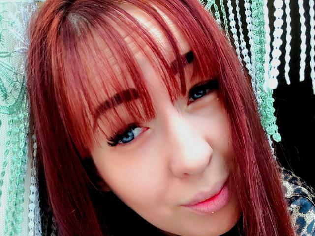 Kohka has red hair, blue eyes, and is smiling with a slight pout. Her eyelashes are long, and she's wearing a leopard print shirt. Her ass is not visible.
