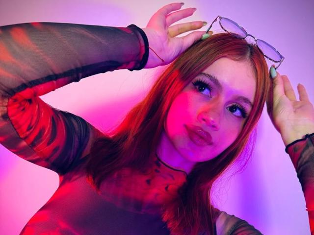 MarilynScotty with red hair, wearing a sheer black top, holding glasses on her head, has pink and purple lighting on her face, looking up with pouty lips.