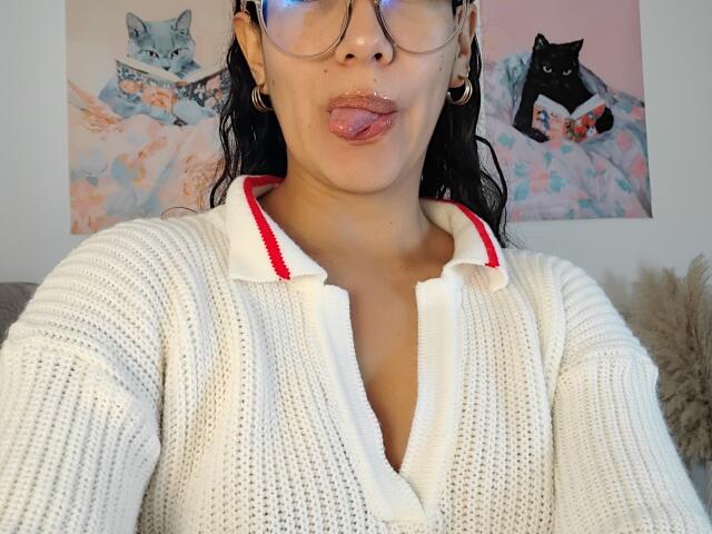 Mllevv with glasses, tongue out, wearing a white knit sweater with red trim, showing small tits, brown skin, and black hair, in a selfie.
