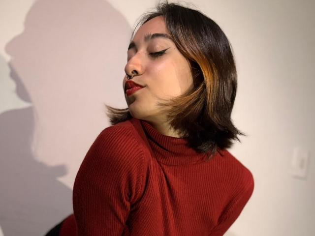 CamiVargas with shoulder-length brown hair, red lipstick, and a red rib-knit turtleneck, eyes closed, shadow on white wall, looking sexy and confident.
