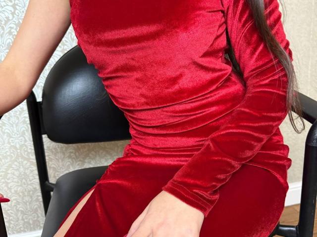 Rouzi's shiny red velvet dress, tight on her ass, clings to her tits and curves, sitting on a black chair with her hand on her thigh.