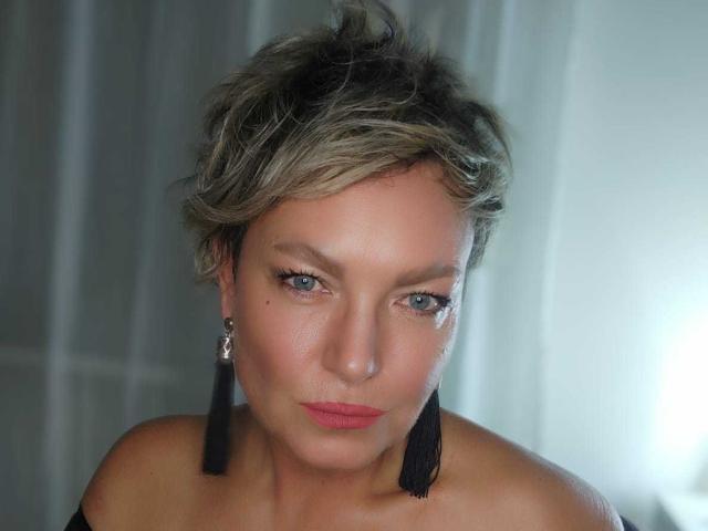 MartaFlow with short, blonde-streaked hair, blue eyes, black tassel earrings, and pink lipstick, staring directly at the camera, showing her bare shoulders and a subtle smile.