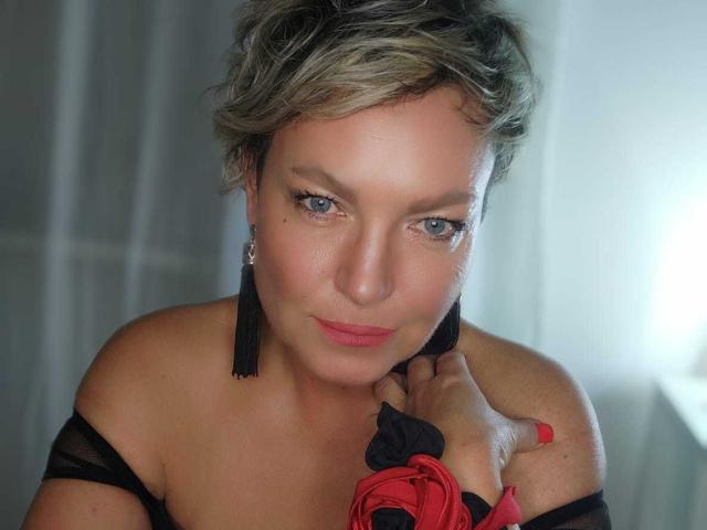 MartaFlow with short, messy blonde hair, blue eyes, and black tassel earrings, holding a red and black fabric against her bare shoulder, smiling softly.
