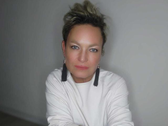 MartaFlow with short, blonde-streaked hair, blue eyes, and pink lips, wearing a white shirt and black tassel earrings, looking slightly to the side.