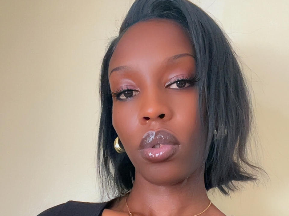 CookieGabby has shiny dark brown skin, glossy pouty lips, long eyelashes, and black shoulder-length hair with a side part. She's wearing gold hoop earrings and a black top.
