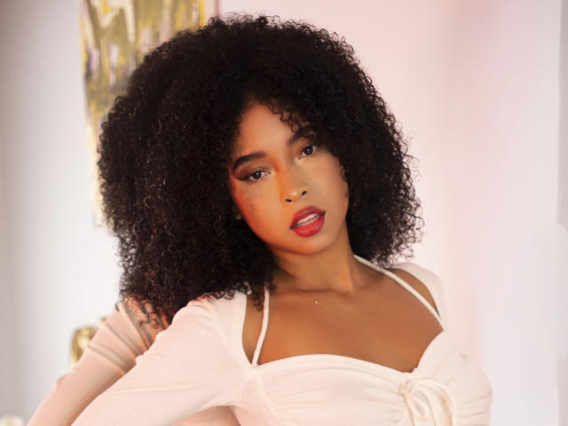 IvyRoberts has curly black hair, red lipstick, and is wearing a white top with a knot. She's looking at the camera with a slight smile. Boobs visible.
