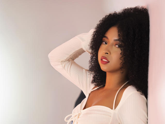 IvyRoberts with curly black hair, wearing a white long-sleeve top, has her hand behind her head, looking to the side with red lipstick and a subtle smile.
