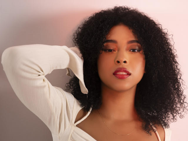 IvyRoberts has curly black hair, brown skin, and red lipstick. She's wearing a white long-sleeve top, with one arm raised, showing off her shoulder. She's looking at the camera.
