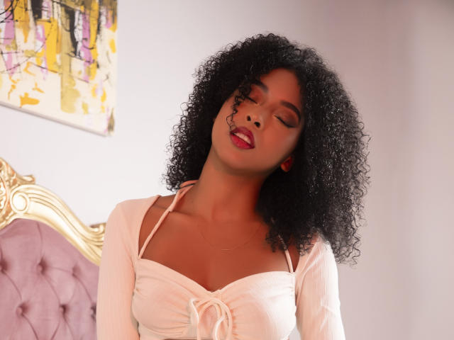IvyRoberts with curly black hair, eyes closed, red lipstick, and white top, head tilted back, showing small tits, in a soft light, sitting on a pink tufted chair.
