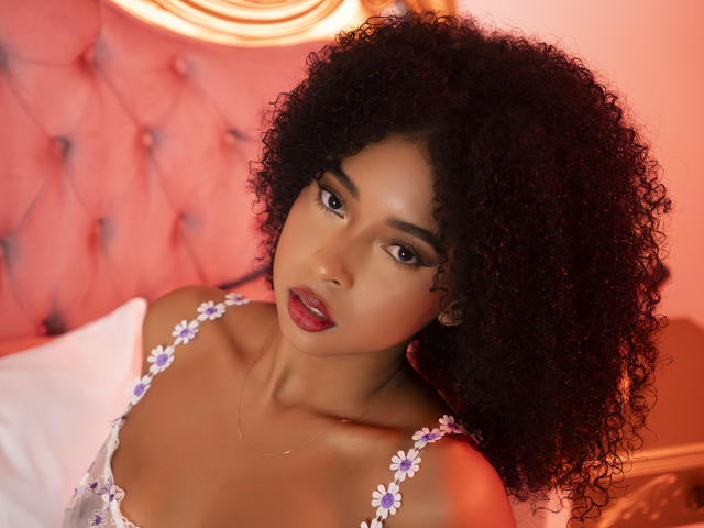 IvyRoberts has dark curly hair, brown eyes, and red lips, wearing a floral top, with a gold necklace, and a sexy, seductive expression. Her tits are visible.