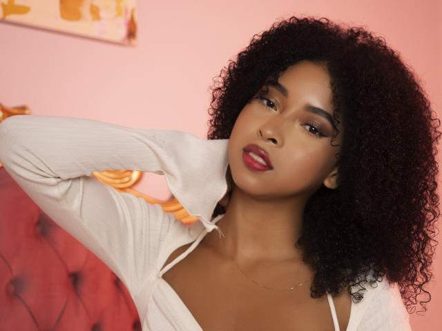 IvyRoberts with curly black hair, wearing a white shirt, has her hand behind her head, showing her red lips and brown eyes, with a subtle necklace. Her tits are visible.