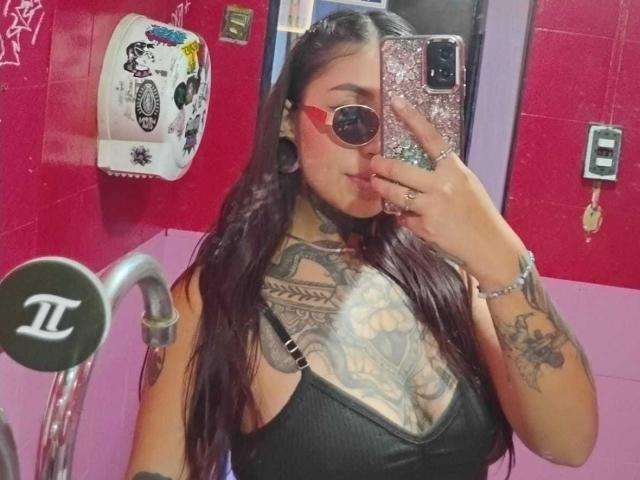 AmberKeller with long dark hair, red glasses, and large tattoos on chest and arm, taking a mirror selfie in a pink bathroom, showing her black tank top and finger.