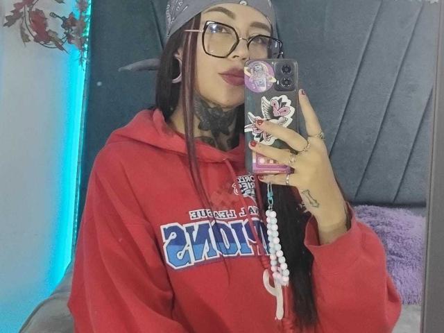 Amber Keller mit langen, schwarzen Haaren, Brille und Tattoos, trägt eine rote Kapuzenjacke und hält ein Handy mit einem Pop-Socket. Sie macht ein Selfie im Spiegel und zeigt eine Perlenkette..