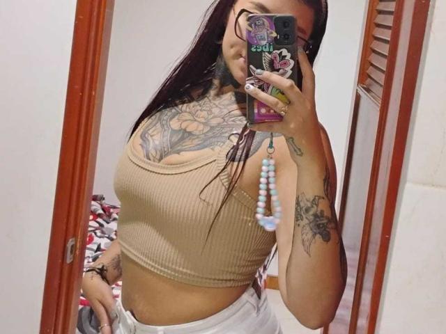 Amber Keller, con tatuajes, vestida con un top corto beige acanalado, pantalones blancos, sosteniendo un teléfono con una funda de colores, mostrando sus pechos y tatuajes en los brazos en un selfie frente al espejo..