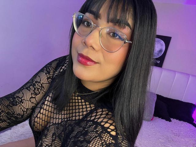 AlexaKiim with black fishnet top, showing her tits through the mesh, wearing glasses, pink lipstick, and black straight hair, smiling at the camera. Bedroom background with purple light.