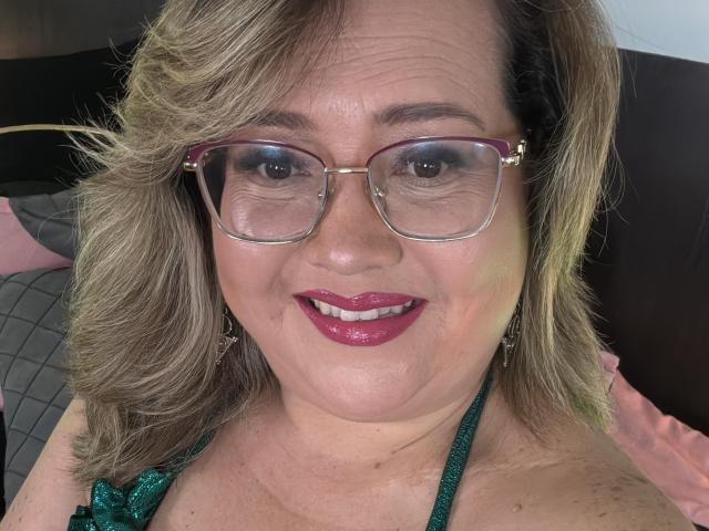 CurvyRosse with glasses, pink lipstick, and blonde hair, smiling at the camera, wearing a green halter top, showing her tits slightly. Her ass is not visible.