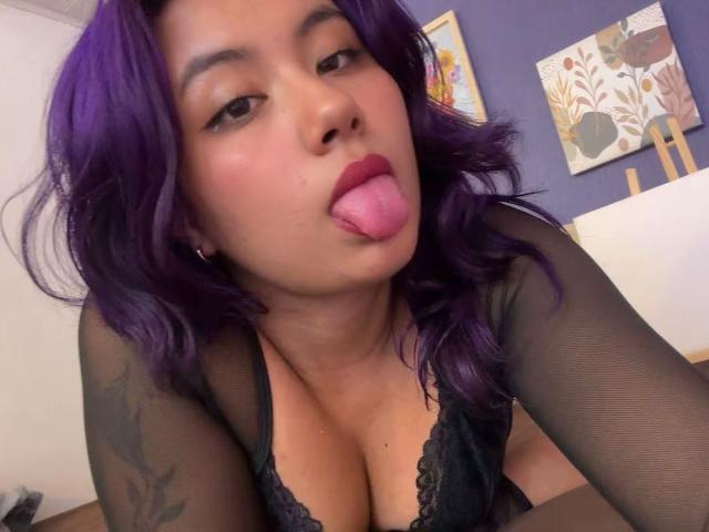 NovaAllen, con cabello morado, un sostén de encaje negro y mangas transparentes, sacando su lengua rosa, mostrando su escote y el tatuaje en su brazo derecho..