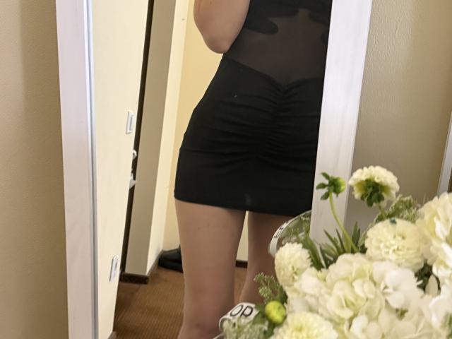 Stefanni's black sheer dress shows her ass and crotch, ruched at the waist, with her bare thighs visible in a mirror reflection, standing next to white flowers.