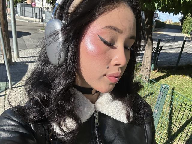 NashiroSakuraii with shiny pink blush, black winged eyeliner, and a black leather jacket with a white fur collar, wearing grey headphones, has a small beauty mark on her cheek.