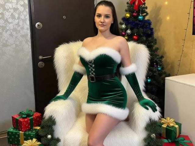 MayaLex vestida con un vestido navideño de terciopelo verde con detalles de piel blanca, guantes verdes, sentada en una silla de piel blanca, con un árbol de Navidad y regalos en el fondo. Pechos, trasero, tetas..