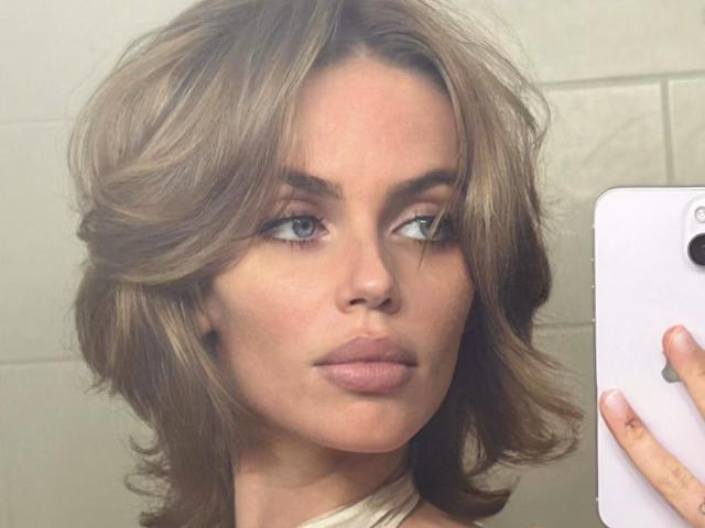 EdenStachurski has wavy brown hair, full lips, and blue eyes, holding a white phone, looking slightly to the side with a neutral expression.