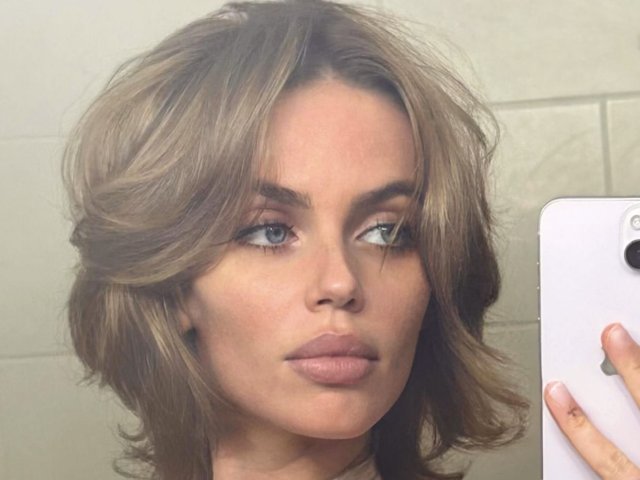 EdenStachurski with wavy, shoulder-length brown hair, full pink lips, and light blue eyes, holding a white phone in a mirror selfie, showing a neutral expression.