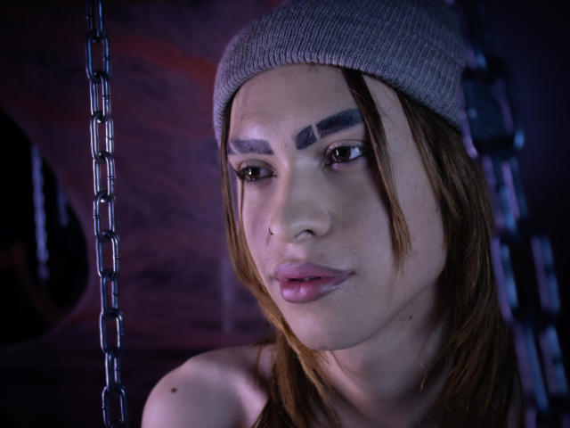 Aaron Finn mit braunen Haaren, der eine graue Mütze trägt, hat dicke Augenbrauen, Eyeliner und pinken Lippenstift, blickt leicht zur Seite und hat einen ernsten Gesichtsausdruck, im Hintergrund ist eine Kette zu sehen..