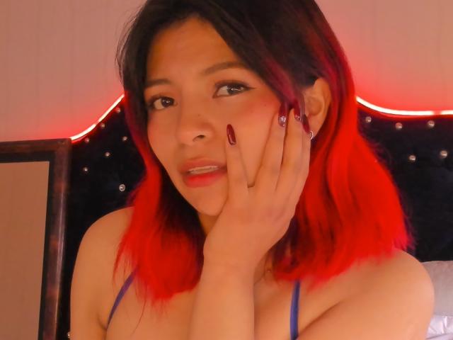 ShofiaWett with red-dyed hair, blue bra, hand on face, red nails, looking at camera, slight smile, soft lighting, bedroom with red LED strip.