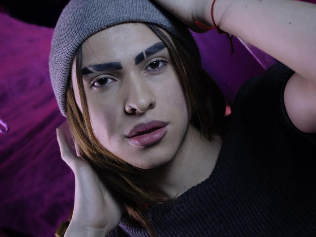 AaronFinn is in a close-up portrait with a neutral expression. He wears a gray beanie and a dark rib-knit shirt. His brown hair is partially covered by the beanie, and he has dark, thick eyebrows drawn on his forehead. AaronFinn's lips are full and slightly parted, with a small nose piercing visible on his right side. His left hand is touching his hair, and a red string bracelet is on his wrist. The lighting is cool, with a pinkish-purple background. The focus is on AaronFinn's facial features and expression.